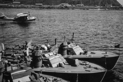 Higgins 78-foot torpedo boats of Motor Torpedo Boat Squadron 13 moored in Attu, Alaska and a PBY Catalina patrol plane taking off in the background, Jul 1943.